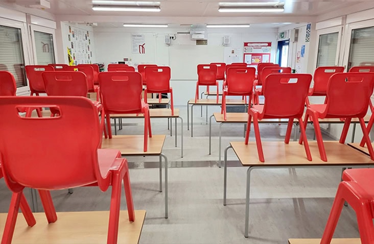 Interior of a modular classroom with rows of tables and stacked red chairs, bright lighting, and educational posters on the walls.