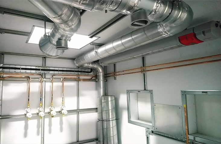 Interior of a plant room showing exposed ducting, pipework and ventilation systems, with copper lines and metal framework visible.
