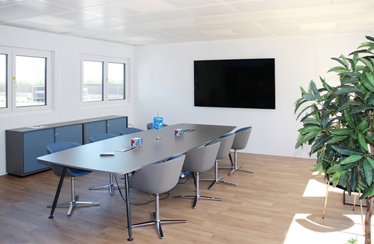Modern modular office meeting space with a sleek table and chairs, a large wall-mounted screen, wooden flooring, and natural light from multiple windows.