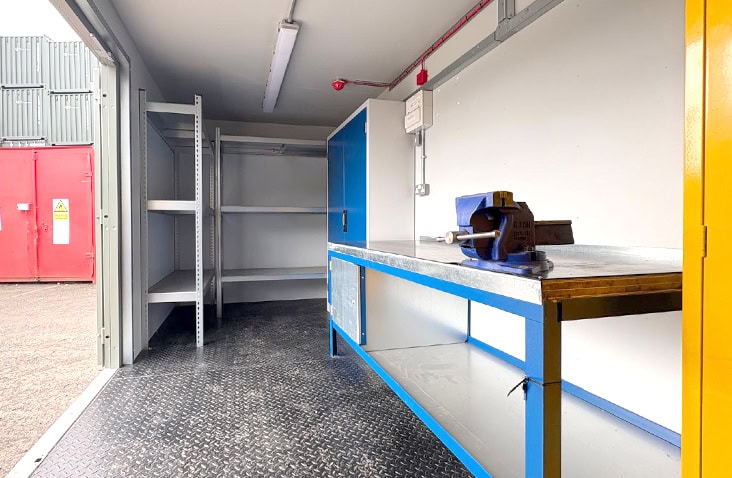 Interior of a containerised workshop with a metal workbench and vice, wall-mounted power sockets, steel floor plating, blue storage cabinet, and open shelving near the entrance.