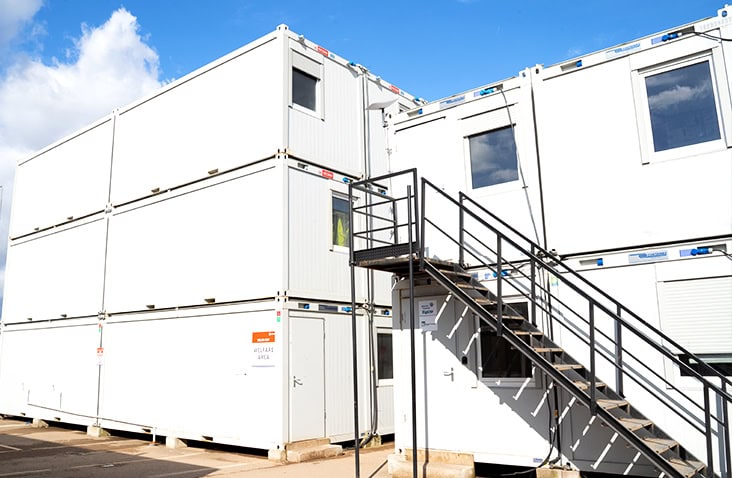 Two-storey modular welfare building made from stacked white units with external metal stair access, located on an industrial site under a blue sky.