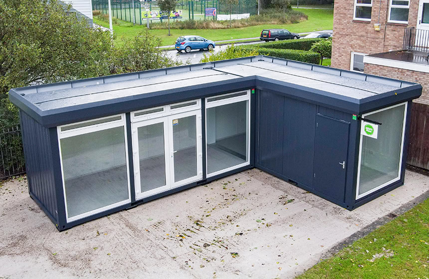 L-shaped modular building with large floor-to-ceiling windows and a dark exterior, installed on a concrete base in a landscaped setting.