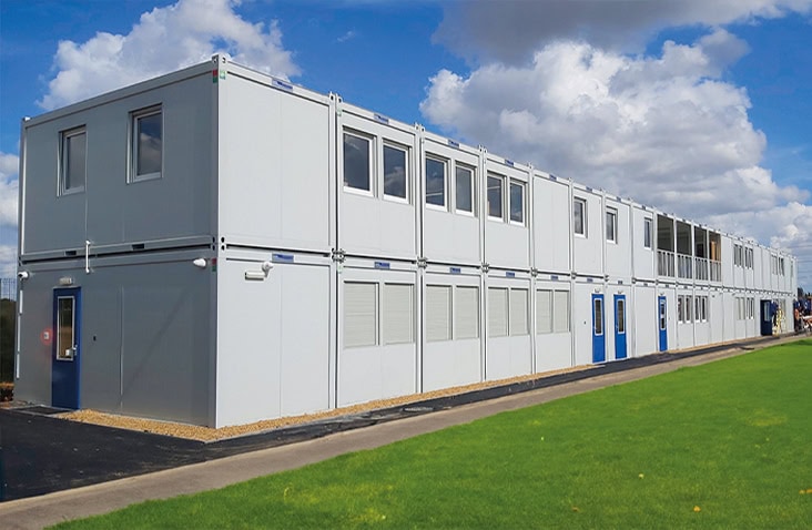 Two-storey modular school building made from stacked white units with blue doors and multiple windows, providing extra learning space beside a grassy area.