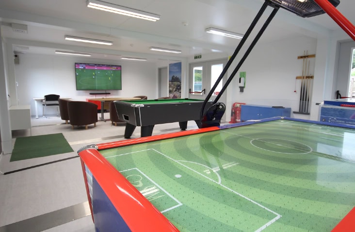 Interior of a modular recreation room featuring a football-themed air hockey table, pool table, seating area with a TV showing a sports match, and wall-mounted games equipment.