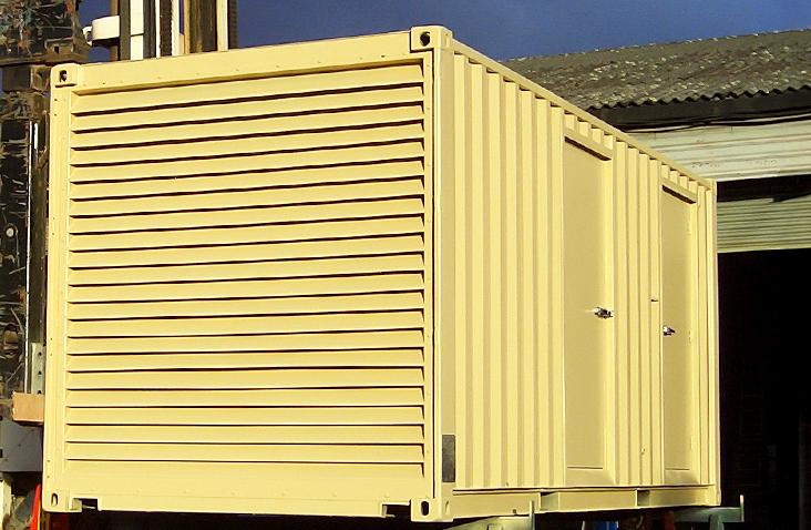 A pale yellow containerised generator enclosure being lifted or positioned by yard equipment. The front end features a large louvred ventilation panel, while the side has two personnel access doors with handles. The unit is made from a converted shipping container by S Jones Conversions and sits outdoors in an industrial yard with buildings visible behind it.