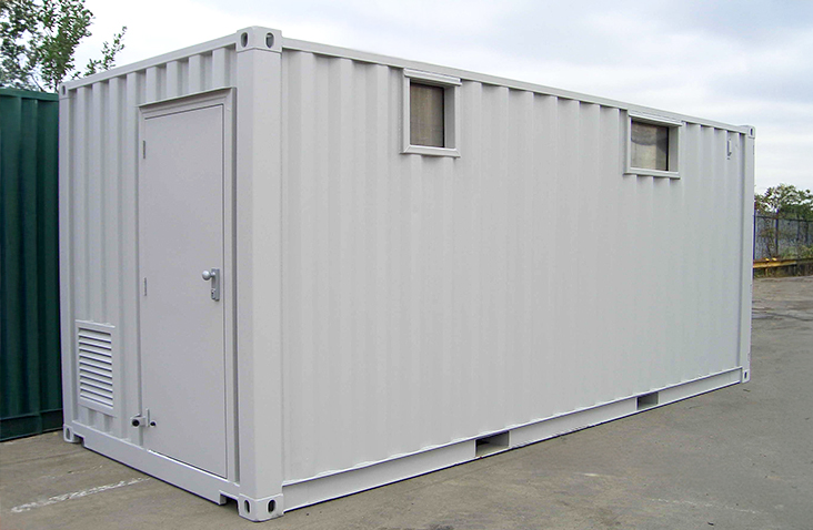 A light grey steel generator enclosure built from a converted shipping container. It has a single personnel access door with a handle on the left side, two small rectangular cutouts near the top of the right sidewall, and a ventilation louvre near the bottom of the door. The enclosure sits on a concrete yard with industrial fencing and trees visible in the background.