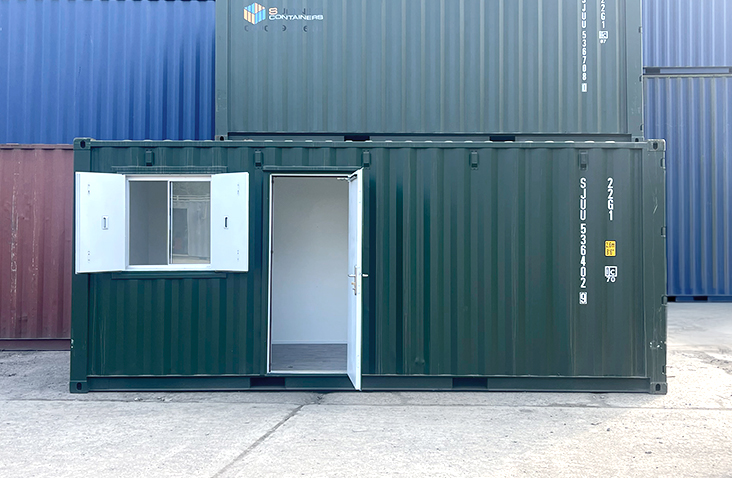 Green 20ft converted shipping container office with a personnel door open and a window fitted with external steel security shutters, positioned in a storage yard with stacked containers in the background.