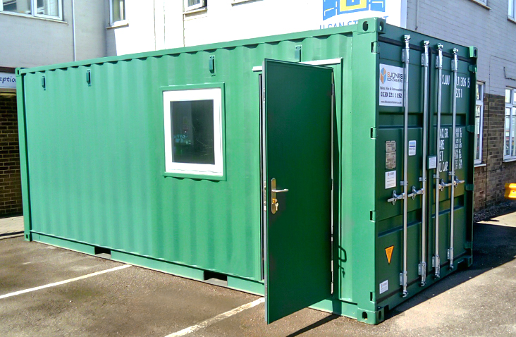 Green 20ft converted shipping container office with a personnel access door open and a single window on the side, positioned in a car park outside a building.