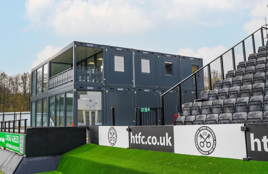Two-storey modular hospitality and retail building beside the spectator stands at Hednesford Town Football Club, featuring large glass panels and an upper viewing balcony.