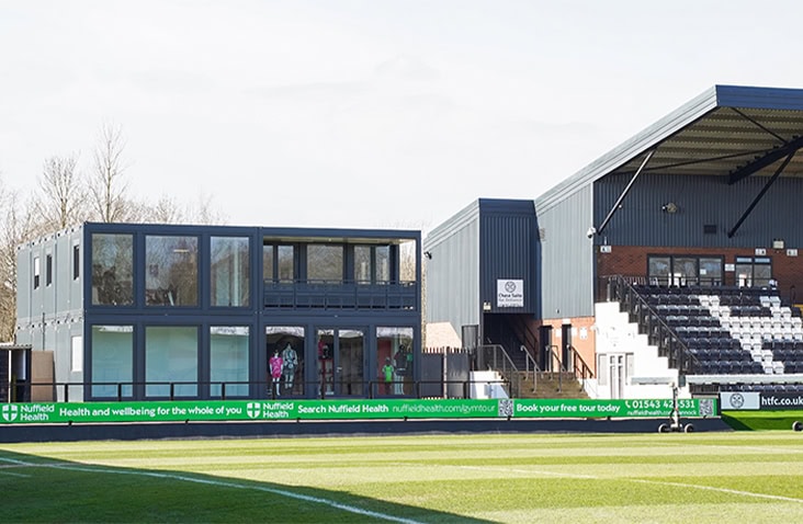 Two-storey modular building with large glass panels installed beside a football pitch, positioned next to the main grandstand at a sports stadium.