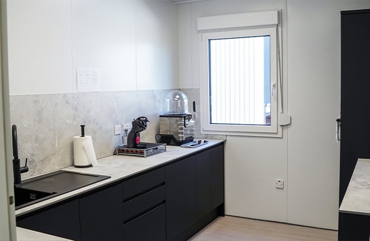 Modern modular kitchen area with black cabinets, marble-effect countertops, a sink, coffee machine, and a window providing natural light.