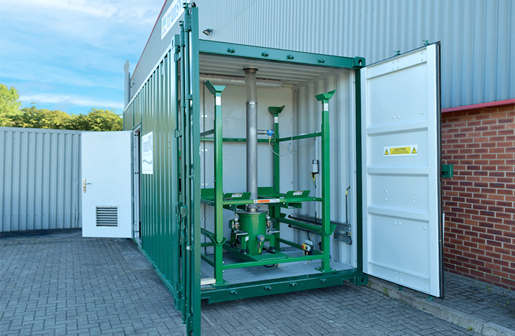 Green converted shipping container housing water treatment equipment, shown with the cargo doors open. The interior contains a steel frame supporting vertical pipework and control components, located on a paved yard beside industrial buildings.