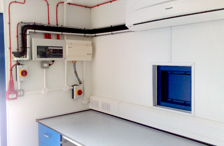 Interior of a converted container fitted out as a small laboratory or control room. The white walls have mounted electrical control panels, conduit, emergency stop buttons, and socket outlets. A countertop with storage sits below, and an air conditioning unit is mounted near the ceiling. A small blue pass-through hatch is built into the wall on the right.