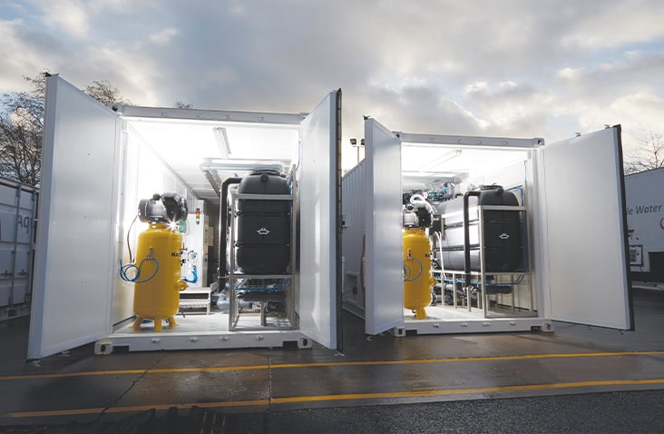 Two white containerised water treatment units with cargo doors open, showing internal filtration equipment including pressurised vessels, pipework, and control components, positioned outdoors on a wet concrete yard.