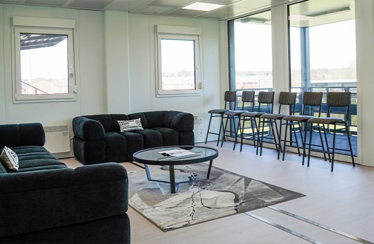 Modern modular lounge area with dark sofas, a coffee table on a patterned rug, and bar seating by large windows overlooking a sports field.