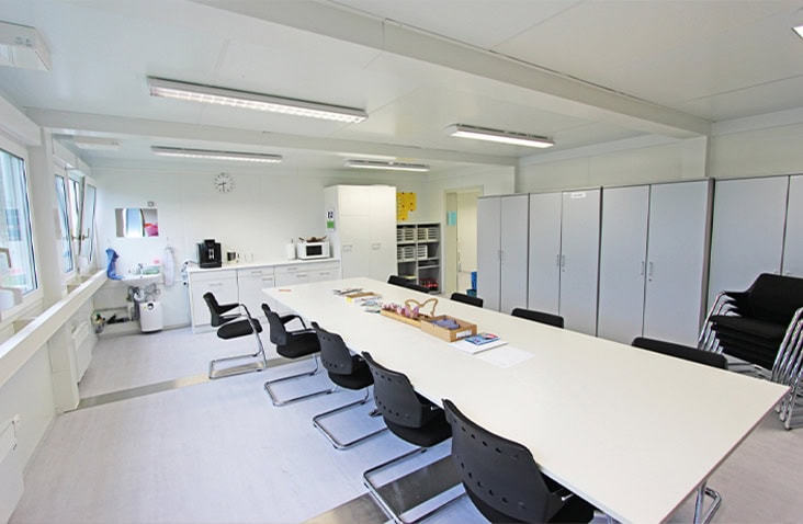 Bright modular office meeting room with a long white table, black chairs, storage cabinets, and a kitchenette area with windows providing natural light.