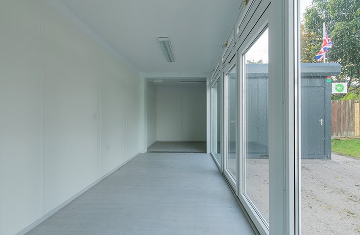 Bright interior of a modular building with white walls, grey flooring, and large windows providing natural light, with another modular unit visible outside.