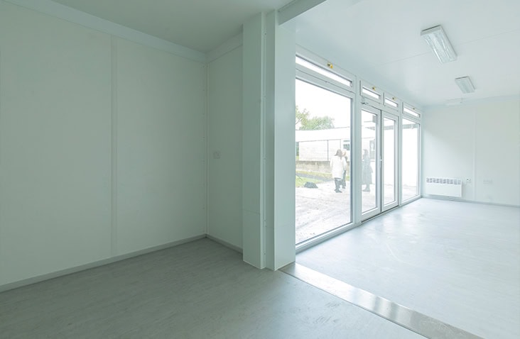 Interior of a modular building with white walls, light grey flooring, and large floor-to-ceiling windows letting in natural light.