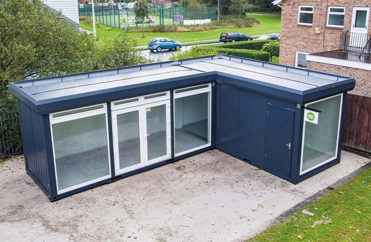 L-shaped dark grey modular building with large glass panels and a single access door, installed on a concrete base beside a brick building and grassy area.