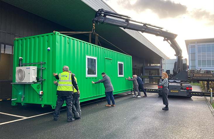 A bright green portable laboratory is being positioned on-site with the help of a crane truck, while several people guide it into place.