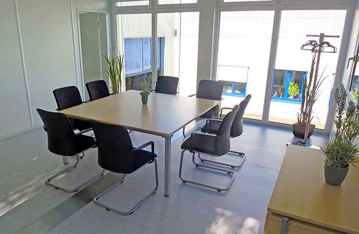 Bright modular office meeting room with large full-length windows, a central table surrounded by black chairs, and indoor plants adding a modern touch.