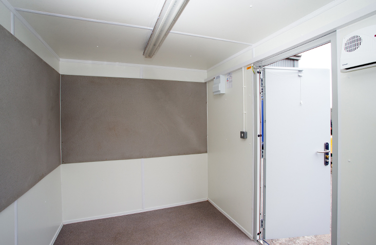 Interior of a converted shipping container office with carpet flooring, white insulated walls, notice boards fitted to the walls, a ceiling light, and an open personnel door leading outside.