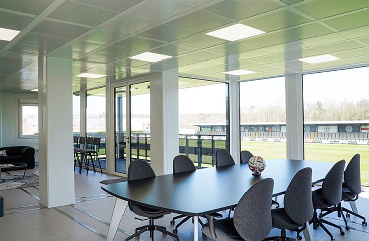 Modern modular office interior with large windows overlooking a sports field, featuring a black meeting table with chairs and an open, bright workspace layout.