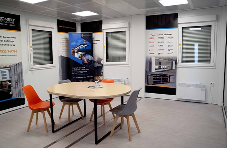 Modular office interior featuring a round meeting table with colourful chairs, wall banners displaying S Jones Containers branding, and windows with shutters.