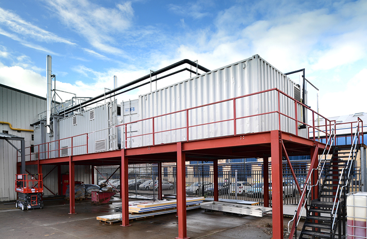 A raised containerised plant room system installed on a red steel platform. The converted white shipping containers house biomass and steam boiler equipment, with external ventilation louvres and pipework running along the roof. The platform has safety railings and an access staircase. The installation is situated on an industrial site with surrounding buildings and fencing visible.