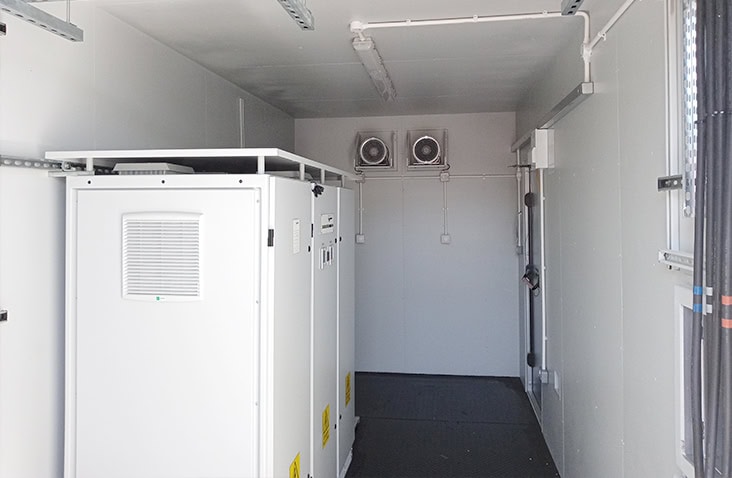 Interior of a containerised switchgear enclosure containing white electrical control cabinets, insulated walls, ceiling lighting, wall-mounted conduits, and a dual-fan ventilation unit at the far end.