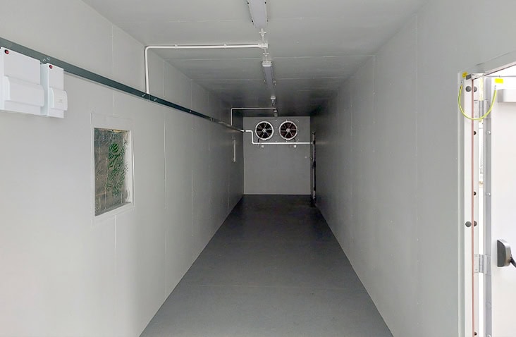 Interior of a switchgear enclosure with insulated grey walls, ceiling lighting, surface-mounted electrical conduits, and a dual-fan ventilation mounted at the far end.