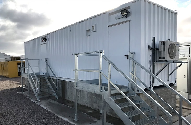 White converted switchgear enclosure elevated on a concrete platform, accessed by metal staircases and railings. The unit features a personnel door, external lighting, ventilation grills, and an external air conditioning unit.