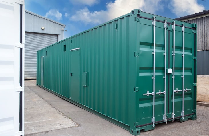 Large green switchgear enclosure with personnel doors and cargo doors closed, positioned in an industrial yard between workshop buildings
