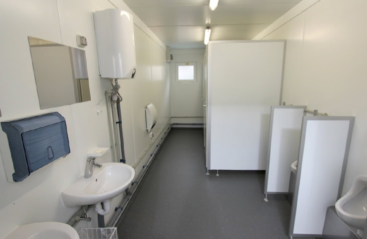 Interior of a modular toilet block with sinks, urinals, cubicles, a wall-mounted water heater, and bright overhead lighting.