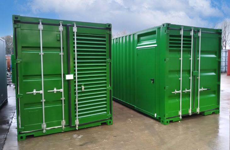 Two green converted switchgear enclosures positioned side by side in an industrial yard. One unit features large louvred ventilation panels on the cargo doors, while the other has solid cargo doors and a personnel access door on the side.
