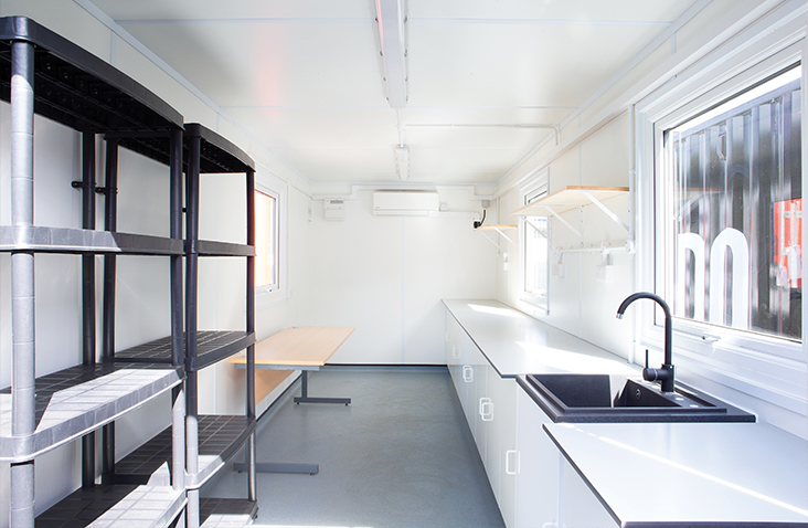 Interior of a containerised research lab with workbenches, shelving units, a sink, bright white walls and windows, designed for practical scientific or operational use.