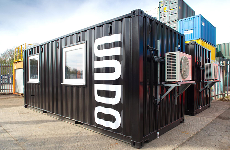 Exterior view of a black containerised research lab with large white ‘UNDO’ branding on the side, fitted with windows and an external air-conditioning unit, situated in an industrial yard. 