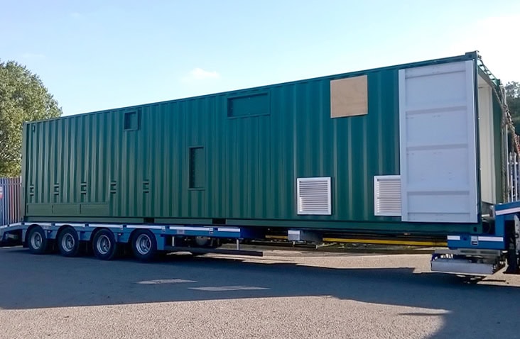 40ft green containerised water treatment enclosure being transported on a flatbed trailer. The unit features side ventilation louvres and access panels, with the cargo doors open at one end.