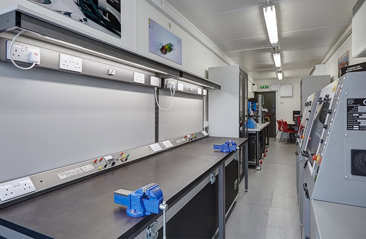 Interior of a containerised workshop with fitted workbenches, vices, electrical sockets, overhead lighting, and industrial equipment along the walls. Seating and additional tools are visible toward the back of the unit.