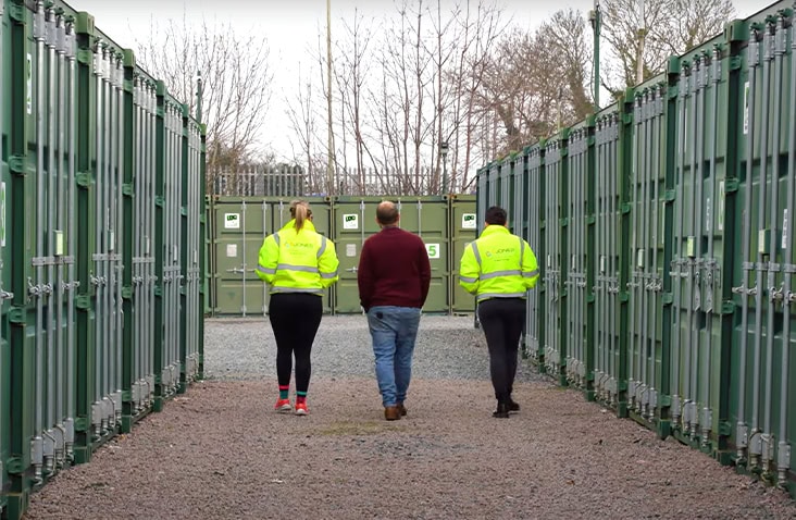 Three people walking down a gravel pathway between rows of green self storage containers on an outdoor storage site, with staff wearing high-visibility jackets. 