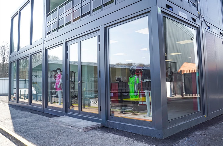 Glass-fronted modular retail building with mannequins visible inside and a double-storey container structure.