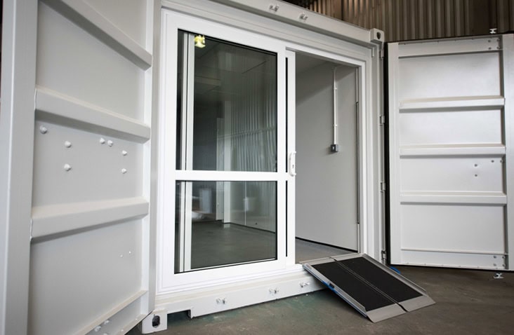 Container conversion with glazed sliding door and access ramp, showing a low-profile ramp leading into a modified shipping container interior with finished flooring and white walls. 