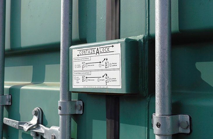 A close-up view of a green shipping container door fitted with a Contain-A-Lock steel lock box. The lock box features an instruction label explaining how to operate the locking system, and the container’s vertical locking bars and metal fixings are visible around it.