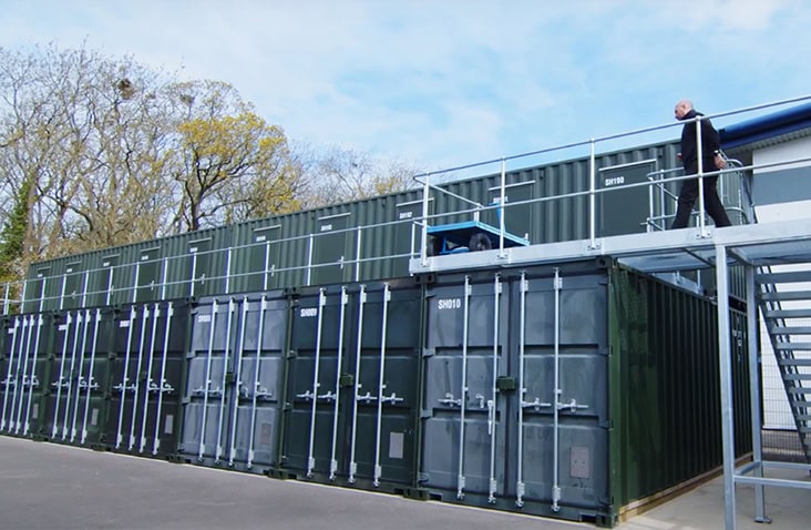 Two-storey self-storage installation made from stacked green shipping containers, complete with an upper walkway and access stairs, with a multi store container sitting atop. 