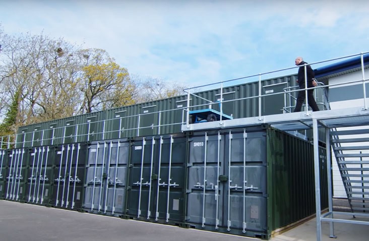 A self storage site featuring stacked 20ft shipping containers with an elevated walkway and steel staircase, with a person walking along the upper level. 
