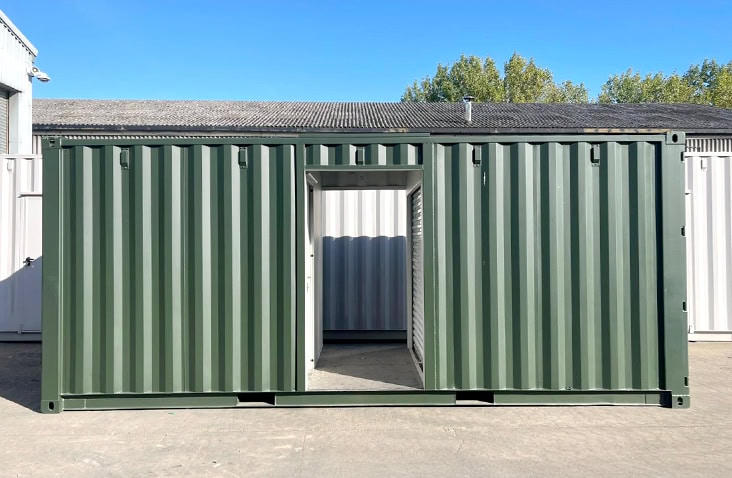 Green steel shipping container (topper unit) with corrugated sides, featuring a central opening that leads to an interior passage. The container is placed outdoors on a concrete surface, with other containers and a building visible in the background under a clear blue sky. 