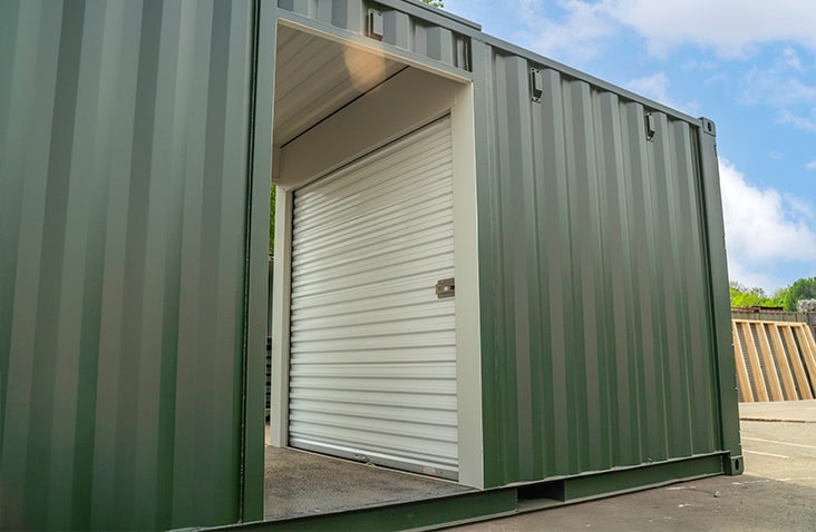 Close-up view of a green steel shipping container with corrugated sides, featuring a topper unit with a central doorway and an internal white roller shutter door. The container is placed on a concrete surface outdoors under a partly cloudy sky. 