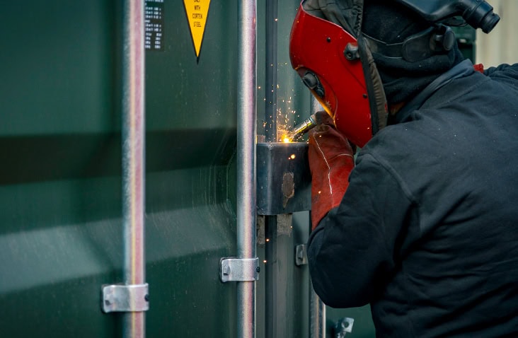 A welder wearing a red welding helmet and protective gloves installs a Contain-A-Lock box onto a green shipping container door. Bright sparks are visible as the lock box is welded into place, with the container’s locking bars running vertically beside the work area.