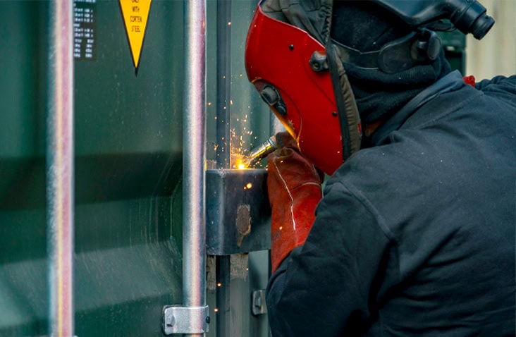     Engineer welding a lockbox onto a green shipping container, with sparks visible during the repair process. 