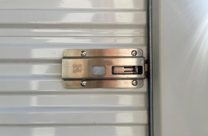 Close-up of a metallic lock mechanism mounted on a white roller shutter door. The lock features a rectangular design with a fingerprint scanner and key slot, secured by four screws on the shutter’s corrugated surface. 